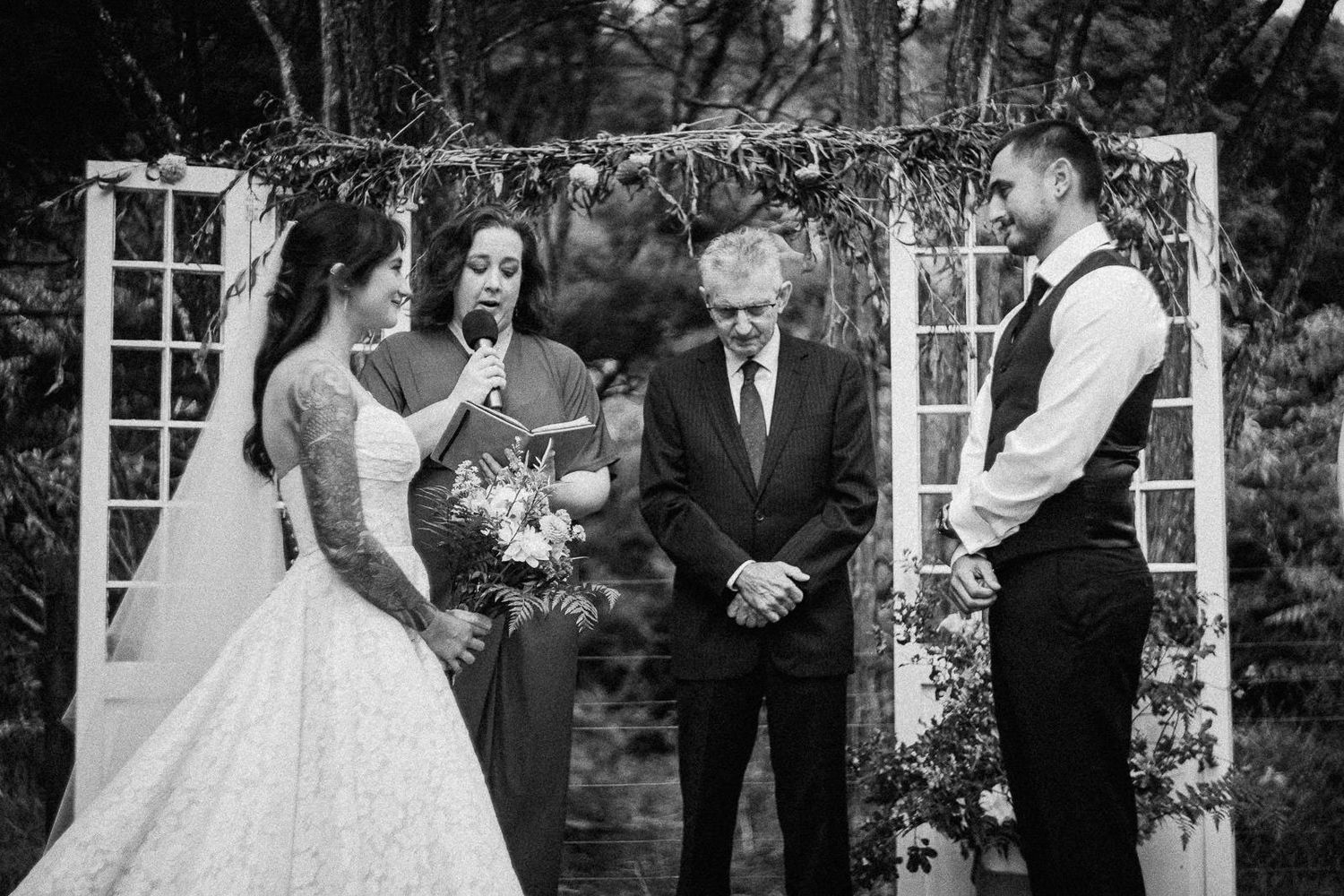 Black and white photo of bride reading vows to groom at outdoor wedding ceremony beneath floral arch.