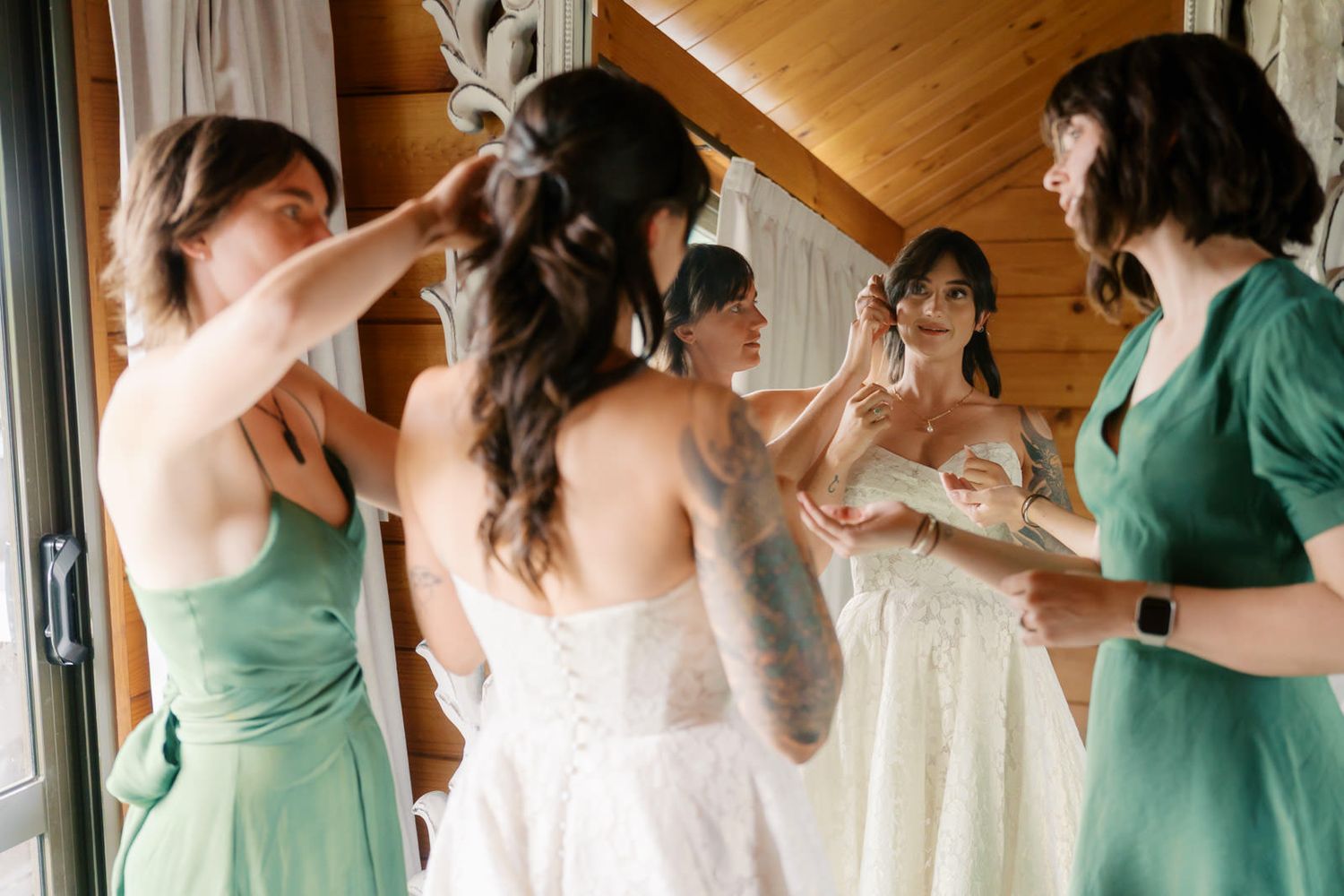 Bridesmaids in green dresses help bride with her white wedding gown in a rustic wooden cabin setting.