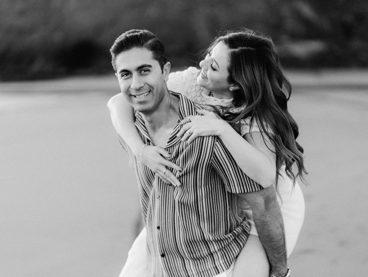 Black and white photo of a couple laughing, woman hugging man from behind on a beach.
