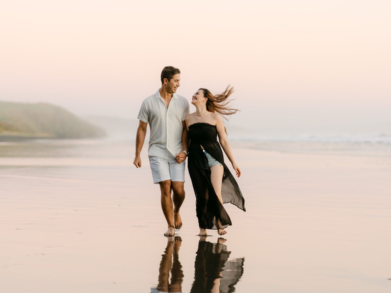 Couple walking hand in hand on a reflective beach at sunset, woman in black dress, man in white shirt and shorts.
