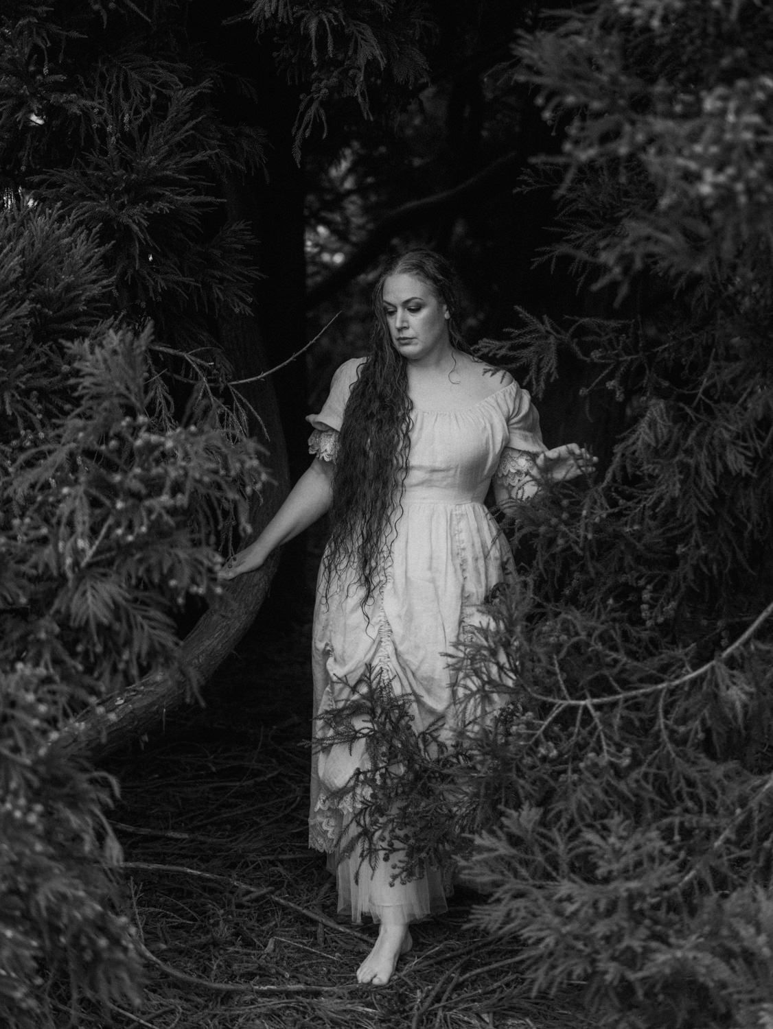 Black and white photo of a woman in a white dress with long hair walking through a dark forest.