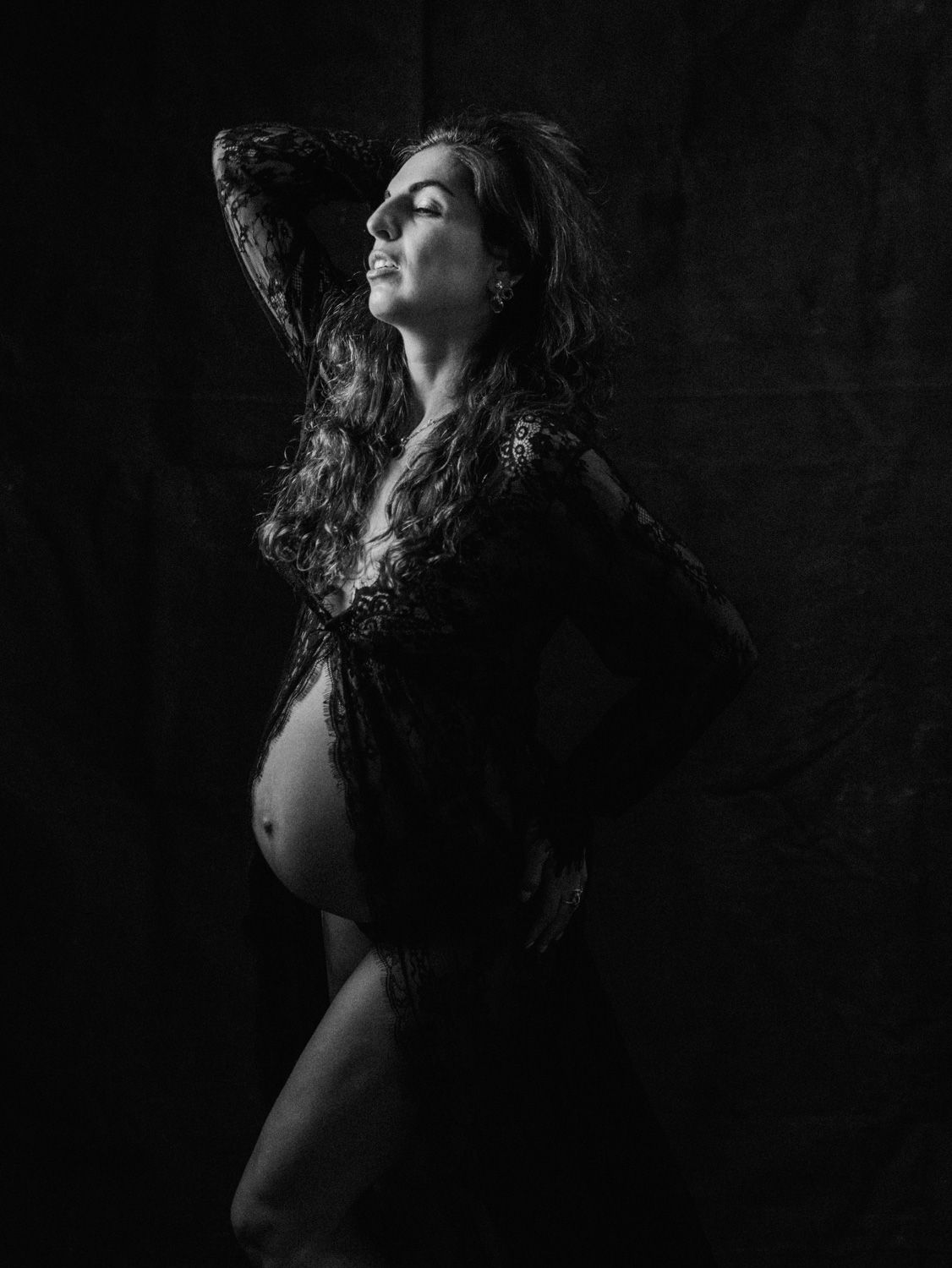 Black and white maternity portrait of a pregnant woman in lace robe posing gracefully against a dark background.