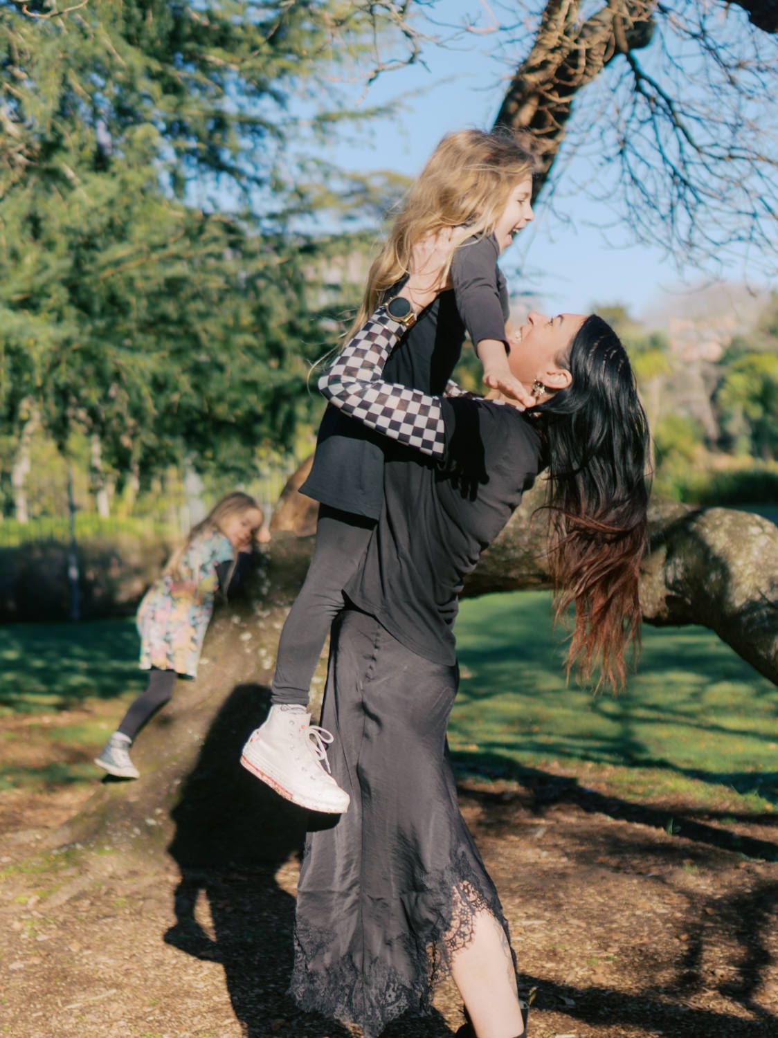 A dark-haired woman lifting a young girl up high while playing in a sunny park.