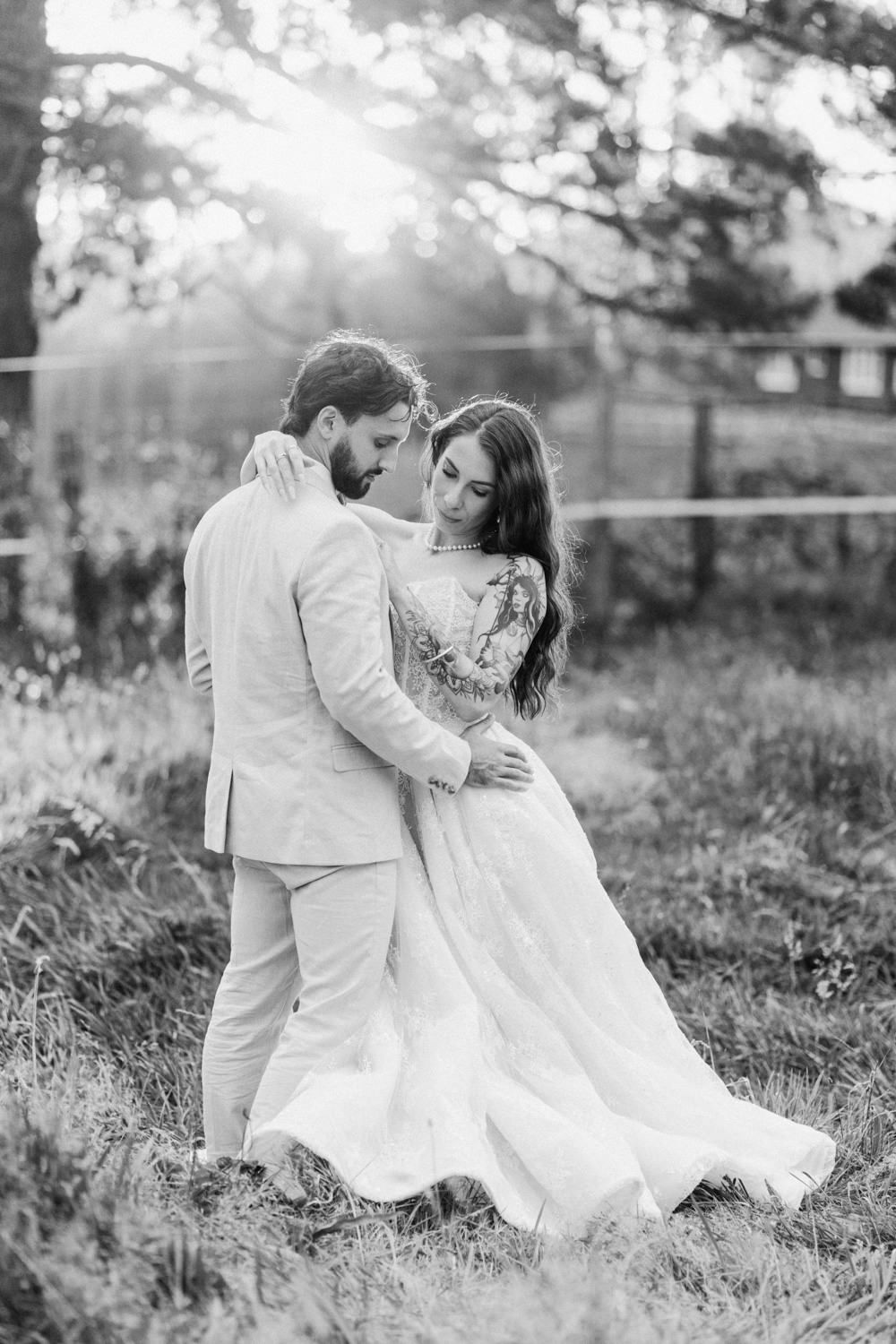 Black and white photo of a bride and groom embracing outdoors at sunset, surrounded by lush greenery.