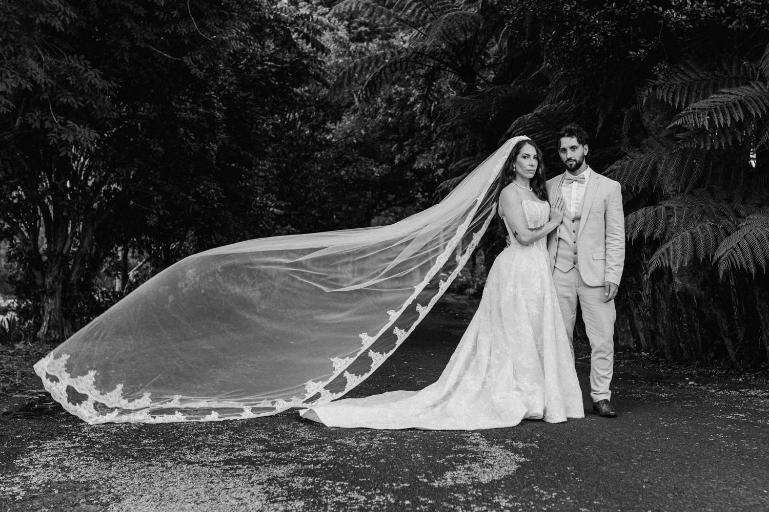 Black and white wedding photo of bride with flowing veil and groom standing together outdoors among lush greenery.