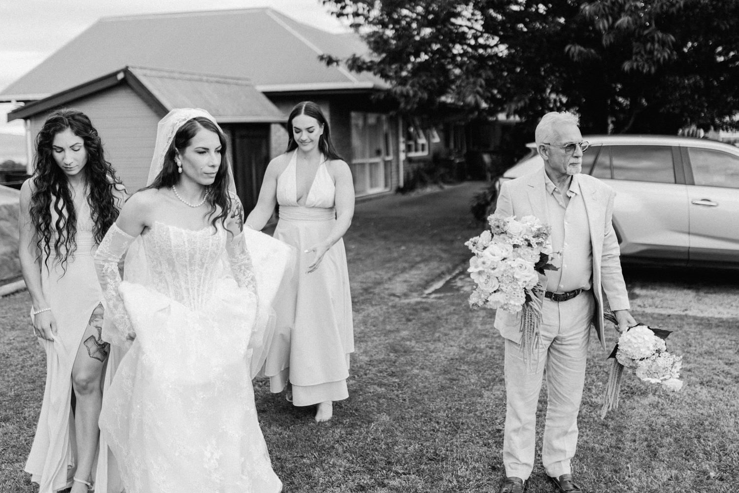 Black and white wedding photo of bride in white gown with bridesmaids and elderly man holding bouquet outdoors.