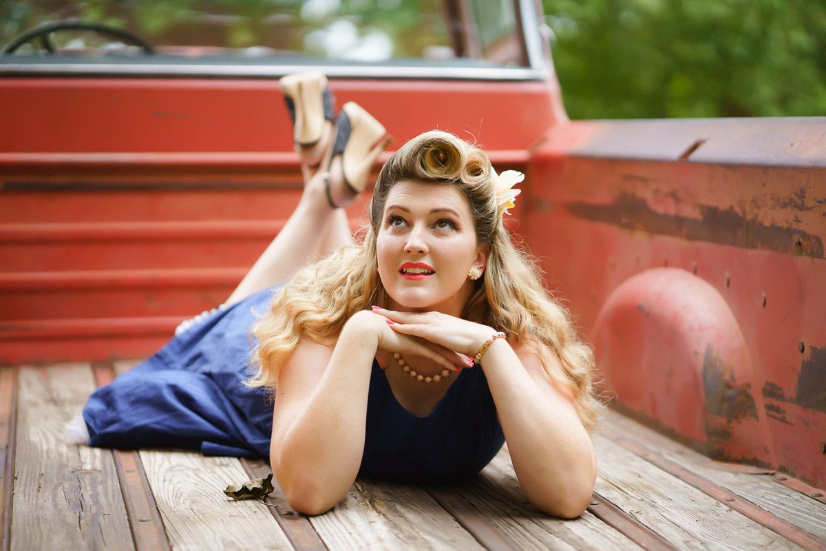Blonde woman in pin-up style lying on wooden truck bed wearing navy dress, posing retro-style near red vintage vehicle.