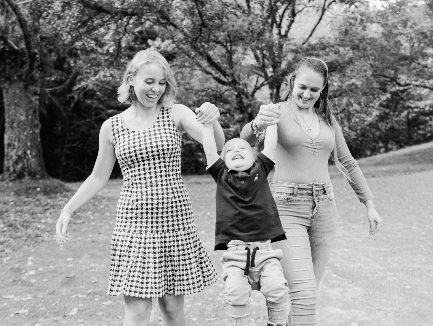 Two women joyfully swing a laughing child between them while walking in a park, captured in black and white.