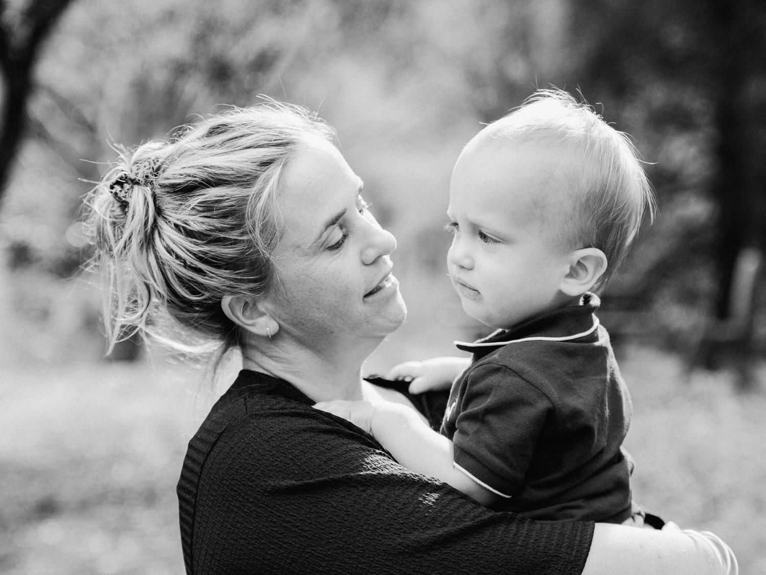 Black and white photo of a smiling blonde woman holding a young toddler boy outdoors in a natural setting.