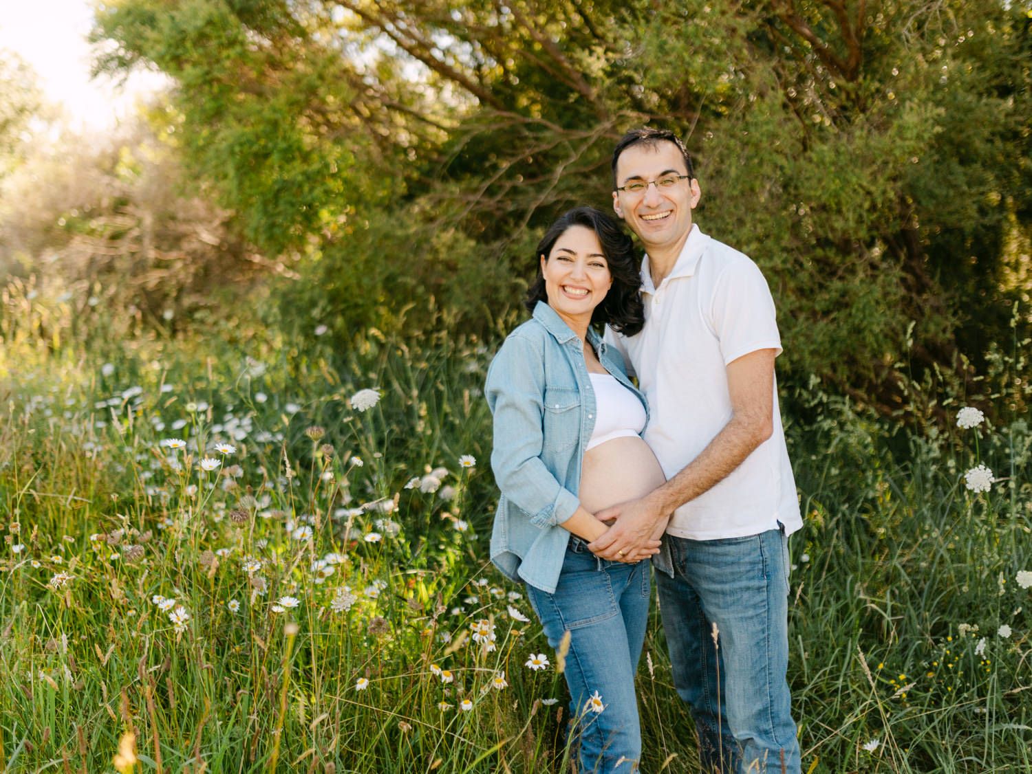 Pregnant couple smiling in a sunny wildflower meadow, woman in denim shirt, man in white polo, cradling baby bump.