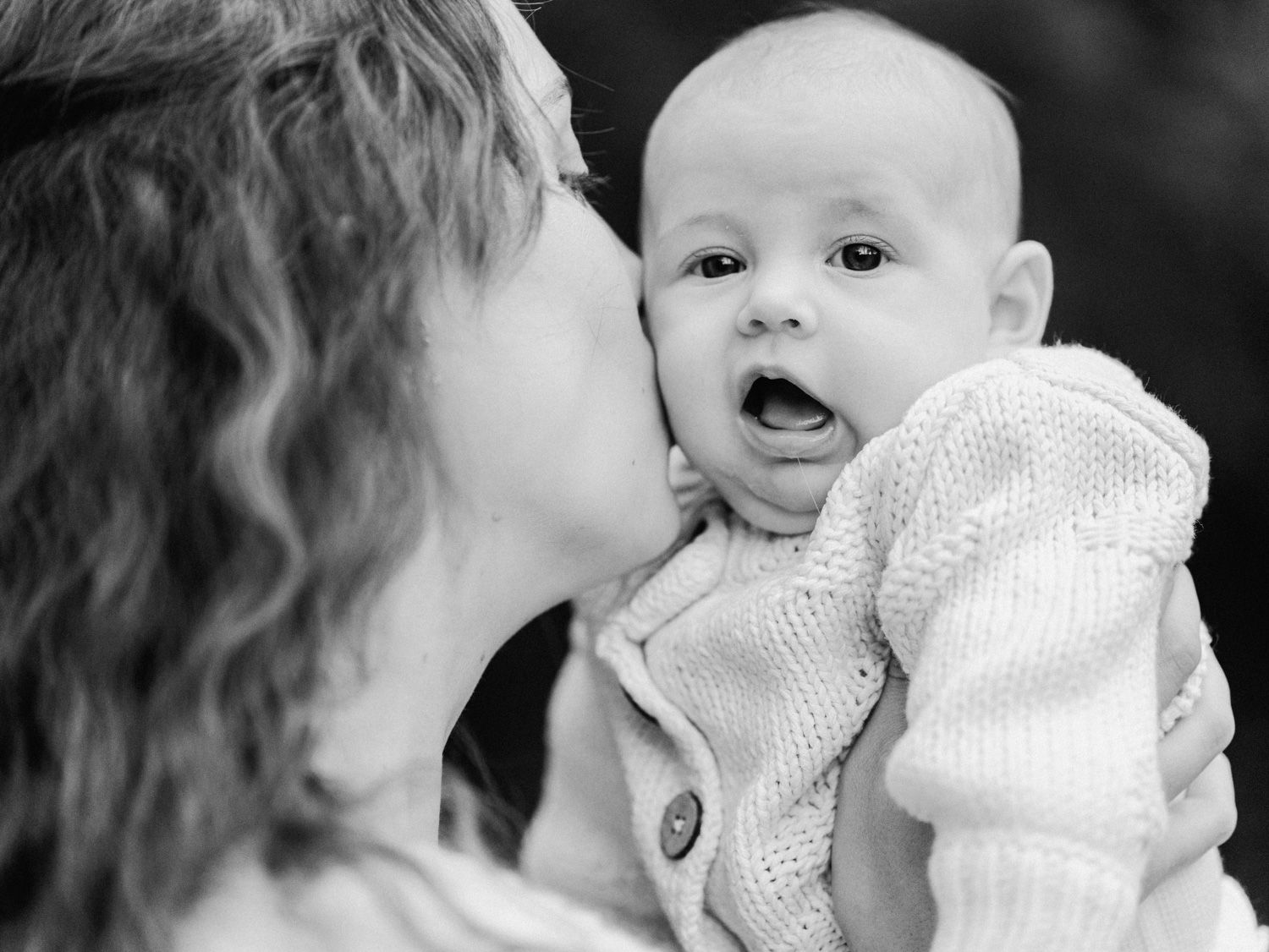 Black and white photo of a mother kissing her wide-eyed baby on the cheek in a tender moment.