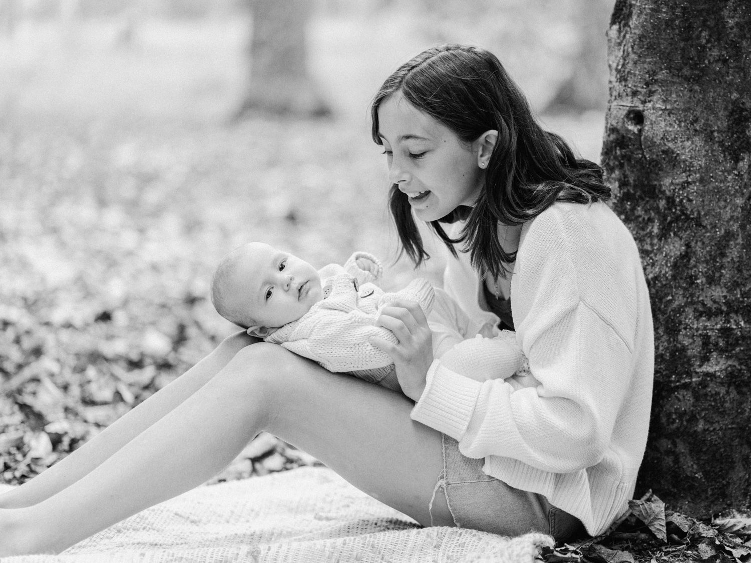 A young mother sits against a tree outdoors, lovingly cradling her newborn baby in a serene black and white photo.