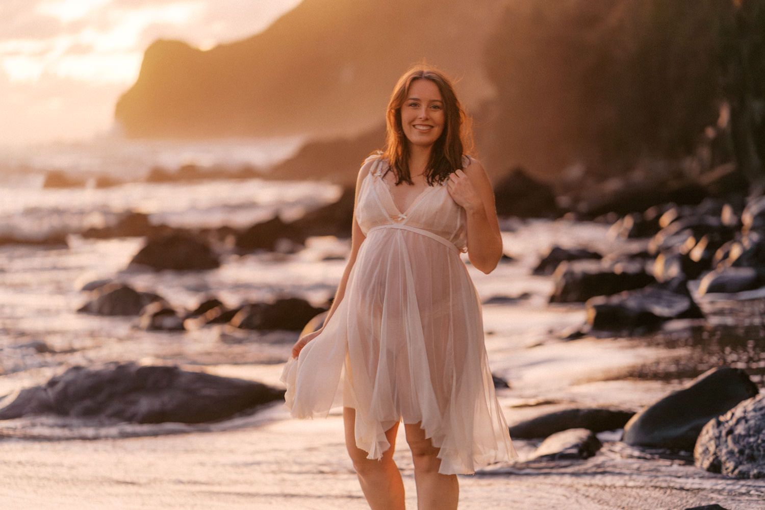 Pregnant woman in white dress on rocky beach at golden sunset.