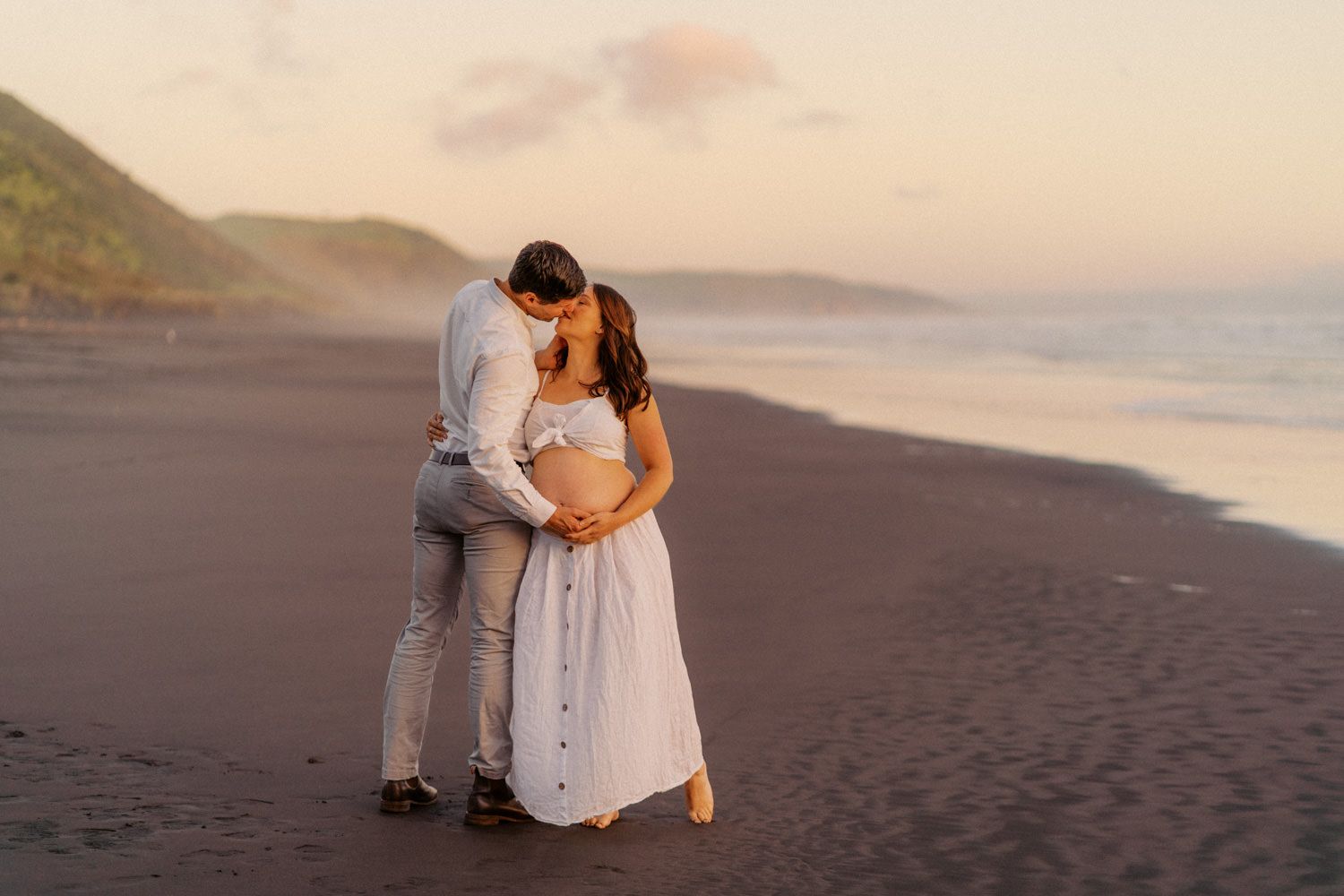 Couple sharing a romantic moment on a black sand beach at sunset, woman in white dress with a baby bump.