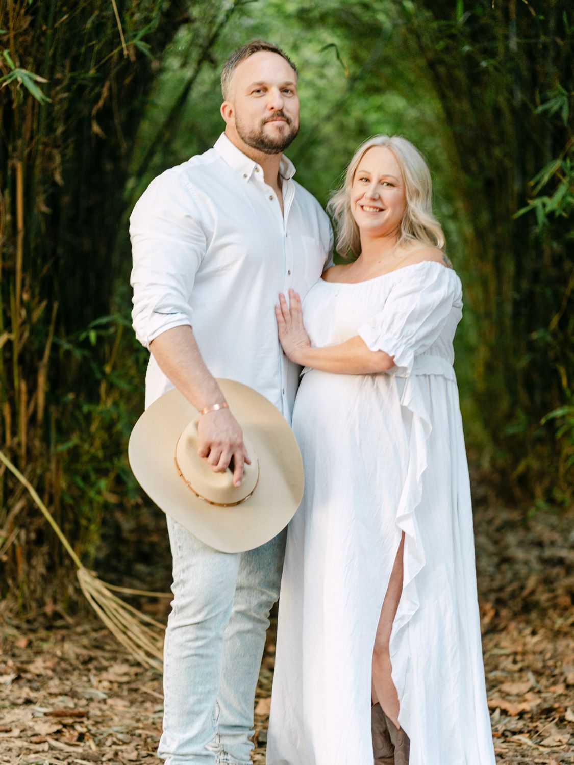 Couple in matching white outfits posing in a bamboo forest, woman in flowing maternity dress, man holding a wide-brim hat.