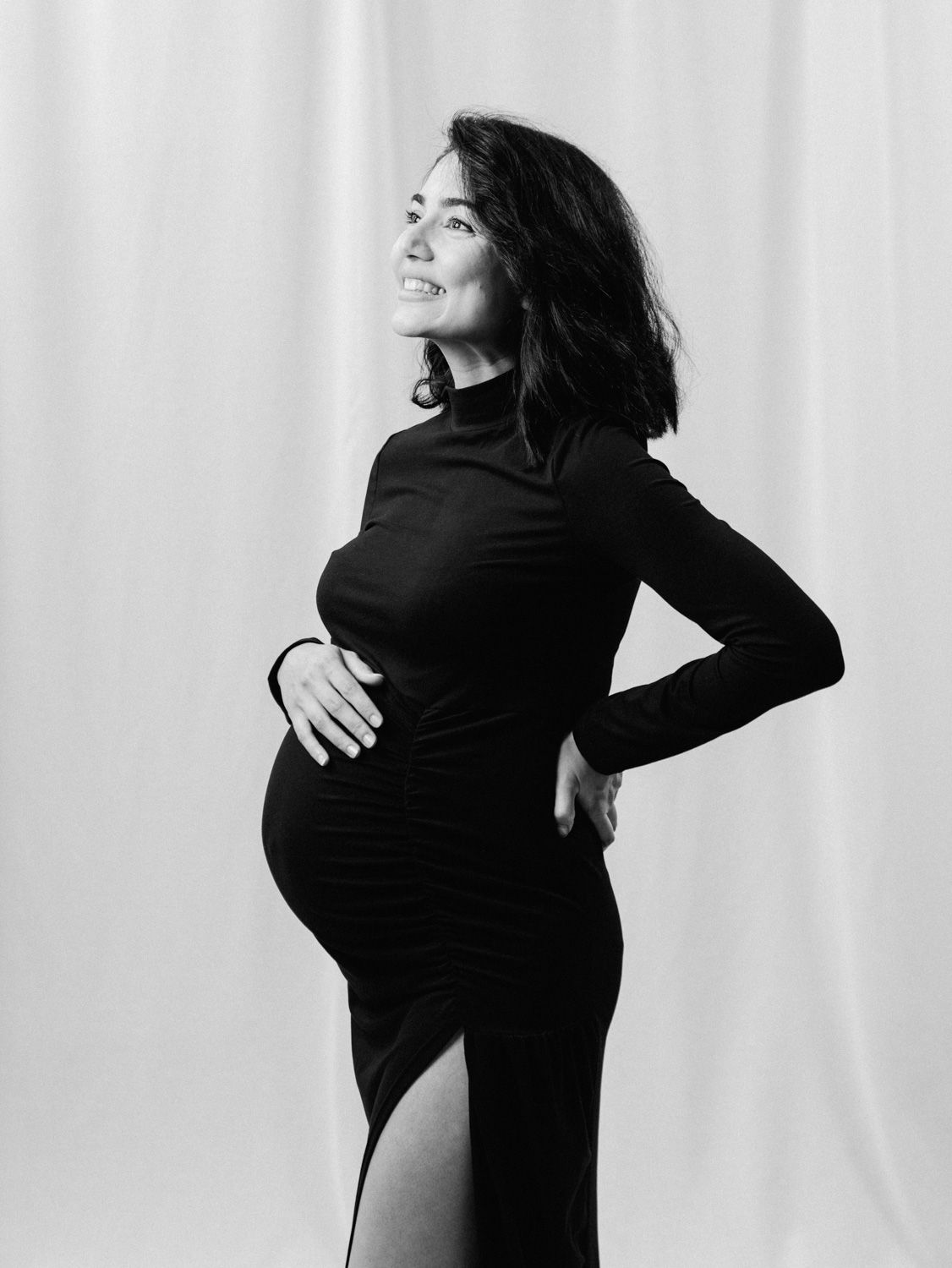 Black and white maternity photo of smiling pregnant woman in elegant black turtleneck dress cradling baby bump.