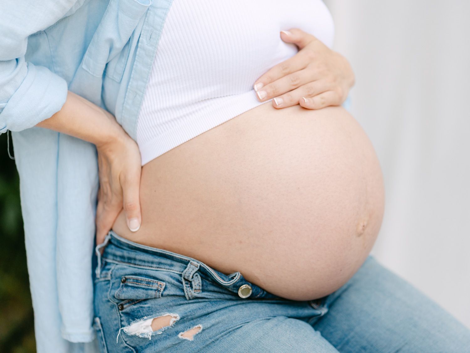 Close-up of a pregnant woman cradling her bare baby bump, wearing a white top and ripped jeans.