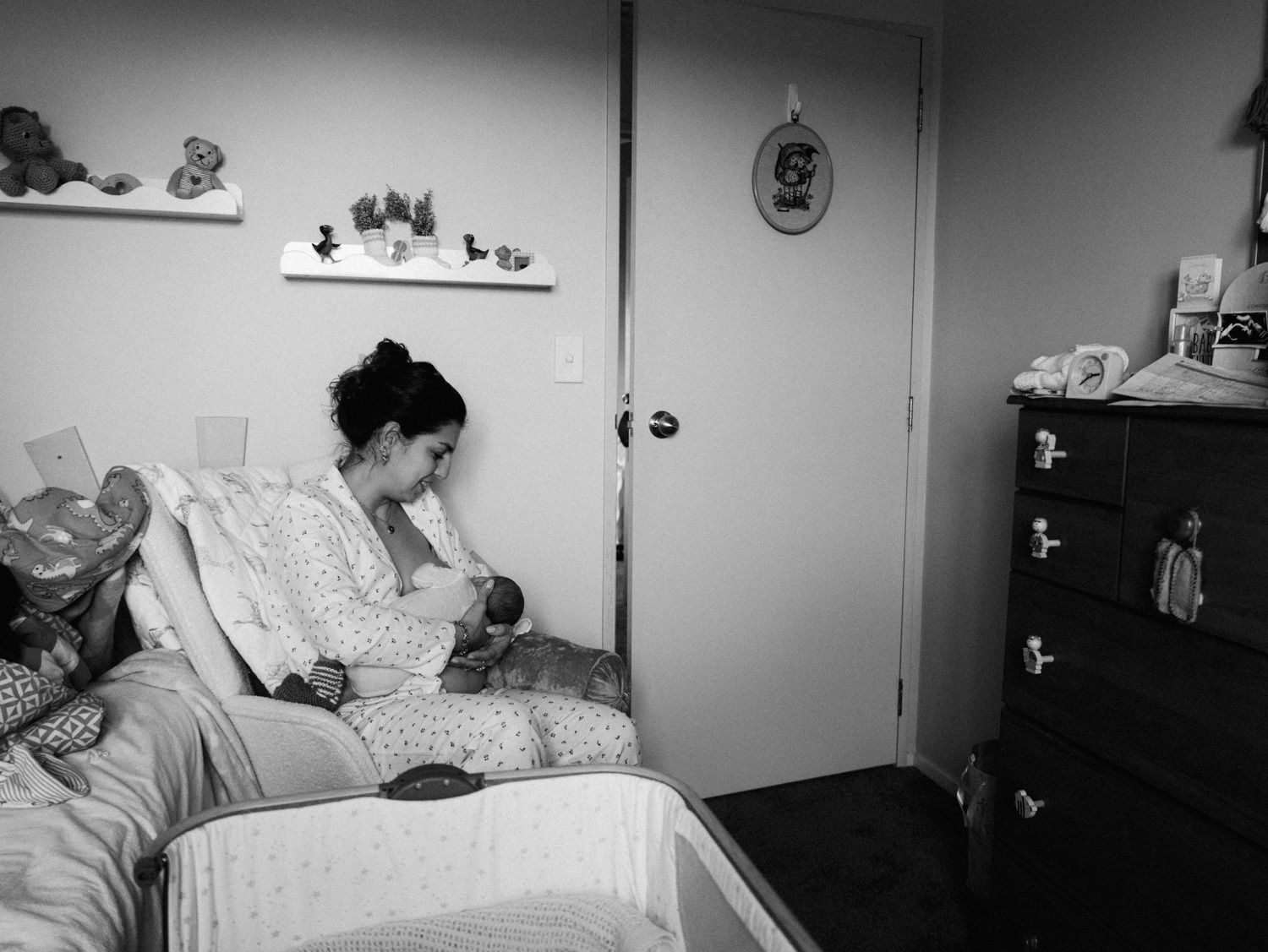 Black and white photo of a new mother sitting on a bed breastfeeding her newborn baby in a cozy bedroom.