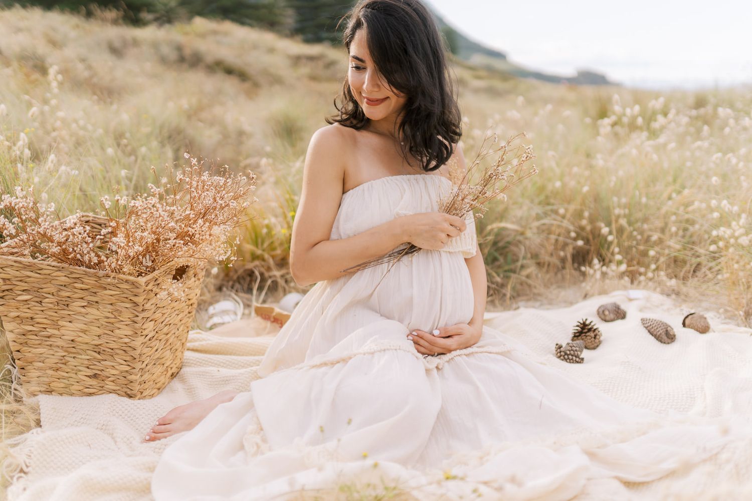 Pregnant woman in white dress sitting in a field holding wildflowers during a serene outdoor maternity photoshoot.