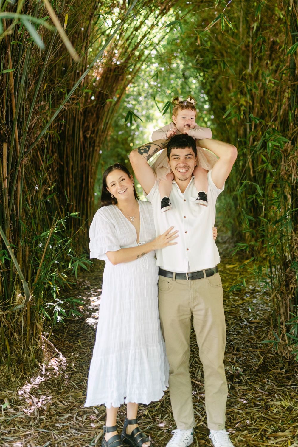 Happy family of three posing in a lush bamboo forest, mother in white dress, father carrying toddler on shoulders.