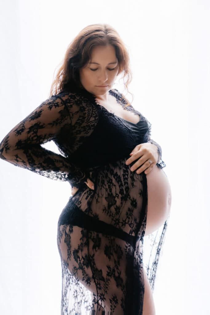 Pregnant woman in sheer black lace maternity gown cradling baby bump against white background.