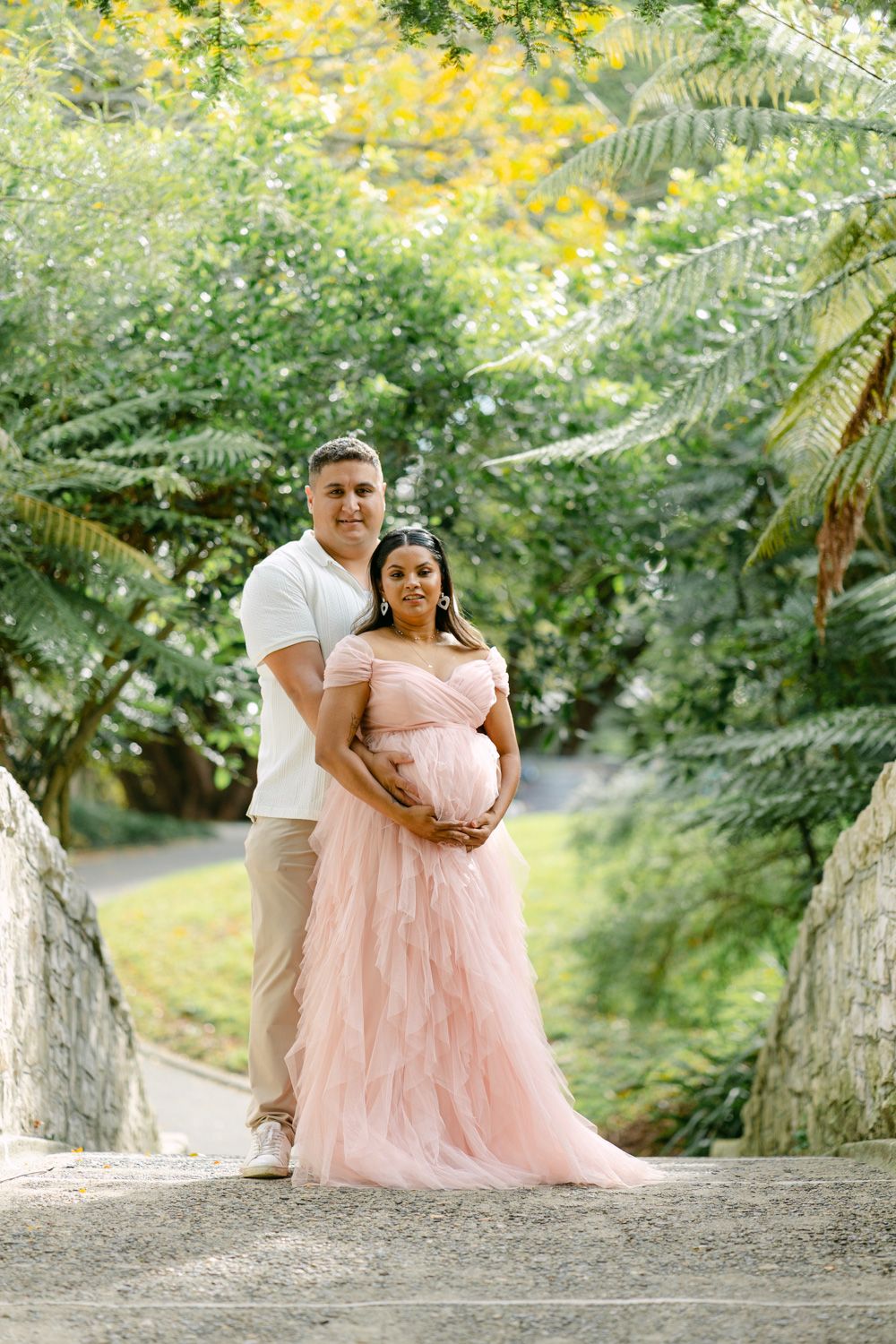 Couple poses for maternity photo in lush garden, woman in pink tulle gown cradling baby bump.