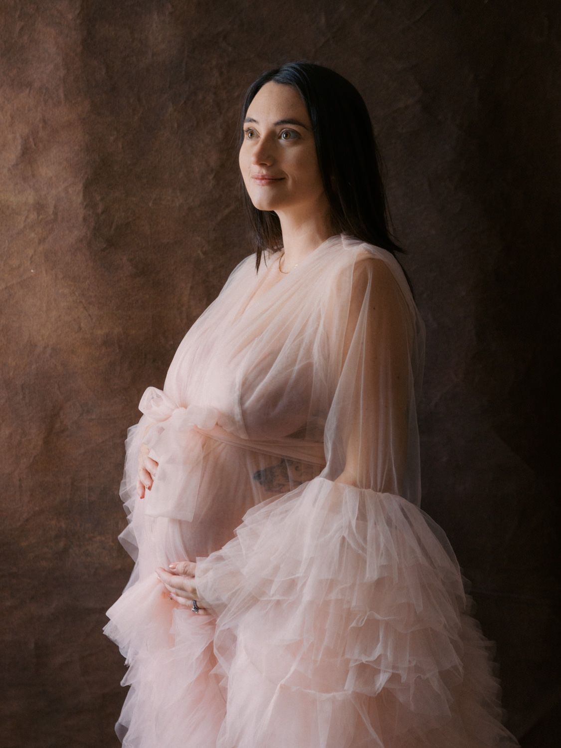 Elegant pregnant woman posing in layered blush tulle maternity dress against dark studio backdrop.