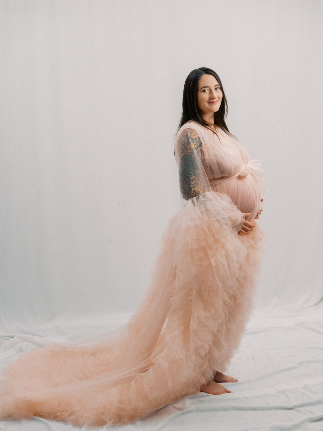 Pregnant woman in a flowing blush pink tulle maternity gown with tattoo sleeve, posing in a studio.