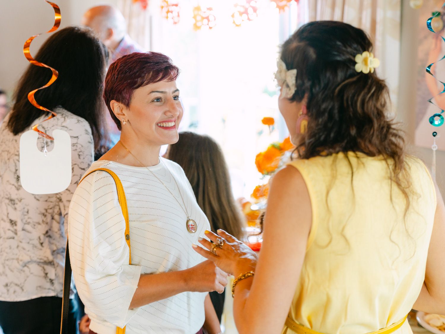 Two women smiling and chatting at a colorful baby shower party with orange and white decorations.