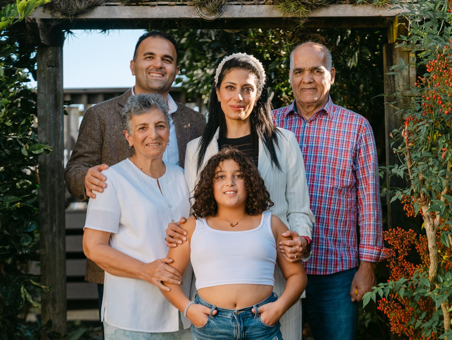 A multigenerational family of five poses together outdoors under a wooden arbor surrounded by lush greenery.