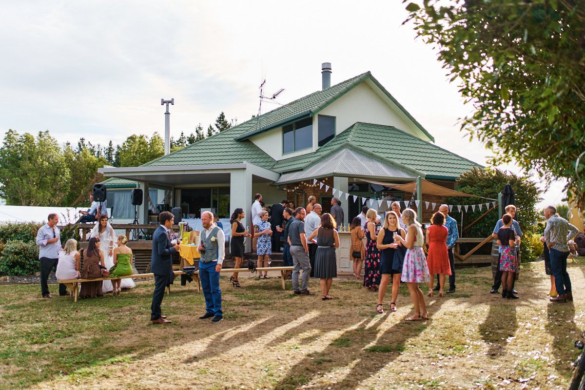 Guests mingling outdoors at a garden party beside a charming green-roofed house on a sunny day.