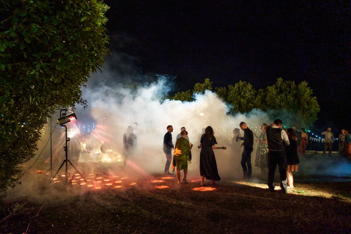 Guests mingle in smoky outdoor night event with colorful fog effects and dramatic lighting among trees.