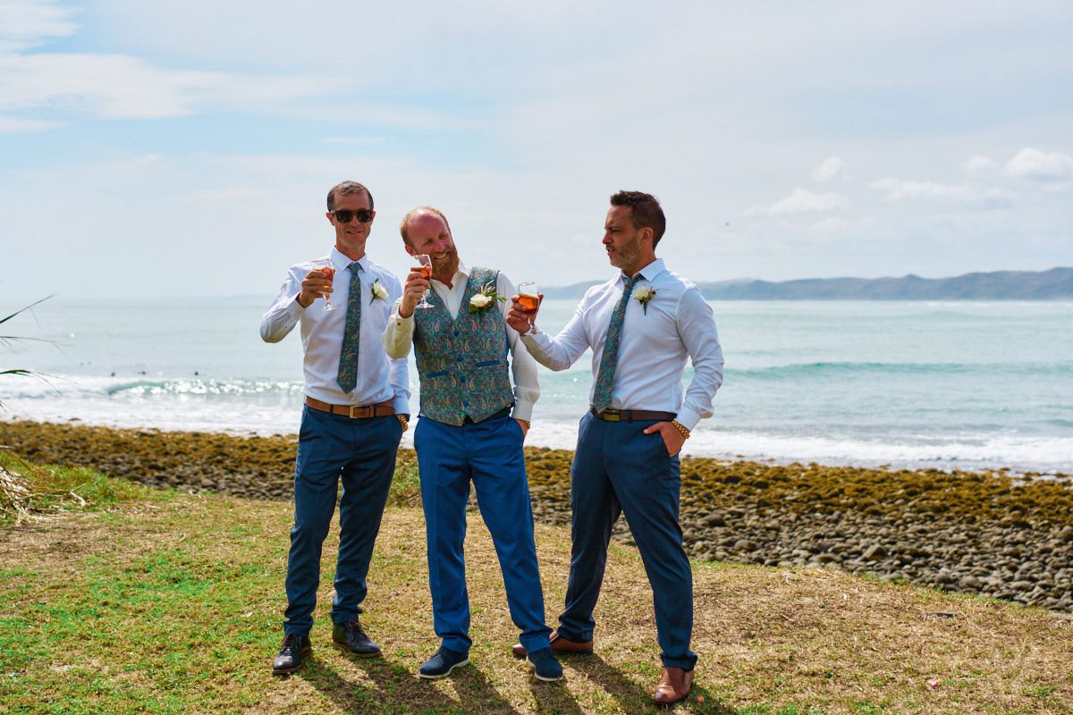 Three groomsmen toasting with drinks on a scenic coastal lawn with ocean waves and mountains in the background.