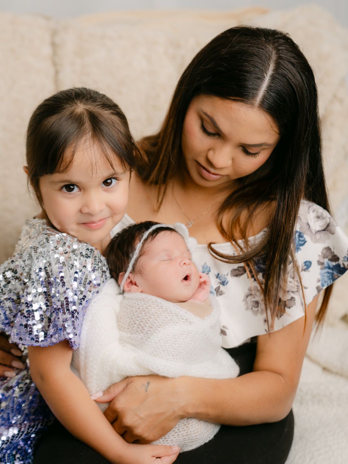 A mother holds her newborn baby while her young daughter in a sequin dress poses beside them on a couch.