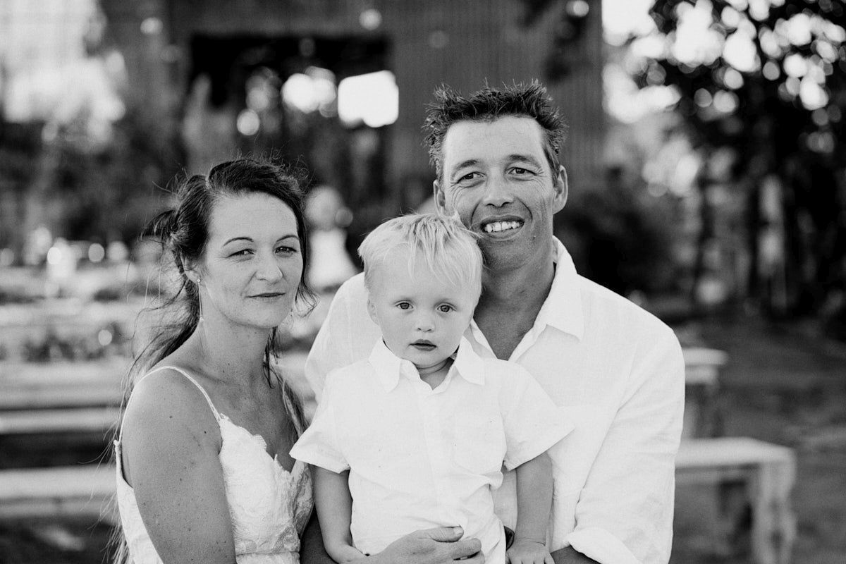 Black and white family portrait of smiling parents with young blond toddler boy outdoors.