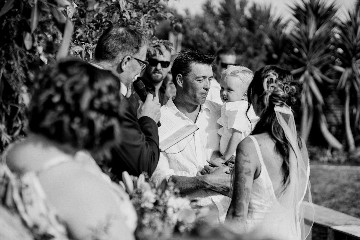 Black and white photo of wedding couple exchanging vows outdoors with guests watching.