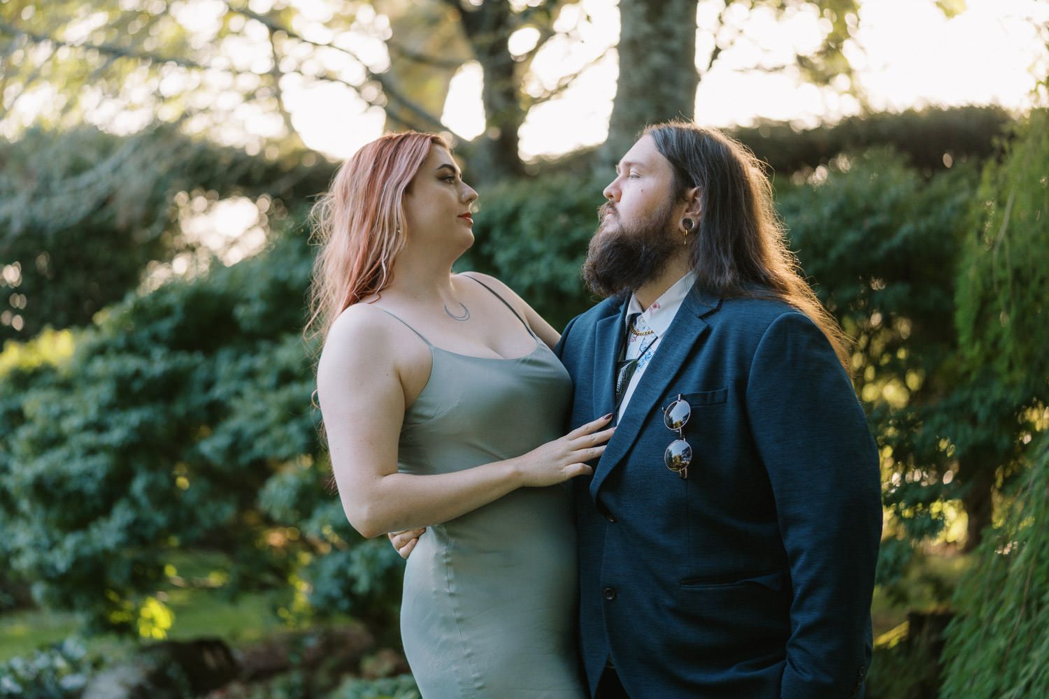 Couple in garden, woman in sage dress with pink hair, man in navy blazer with long hair, sharing a romantic moment.