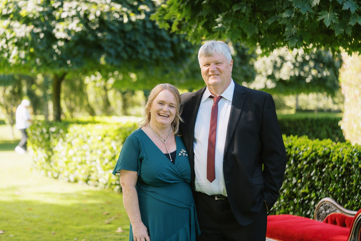 Smiling couple posing outdoors in a lush garden, woman in teal dress and man in suit with red tie.