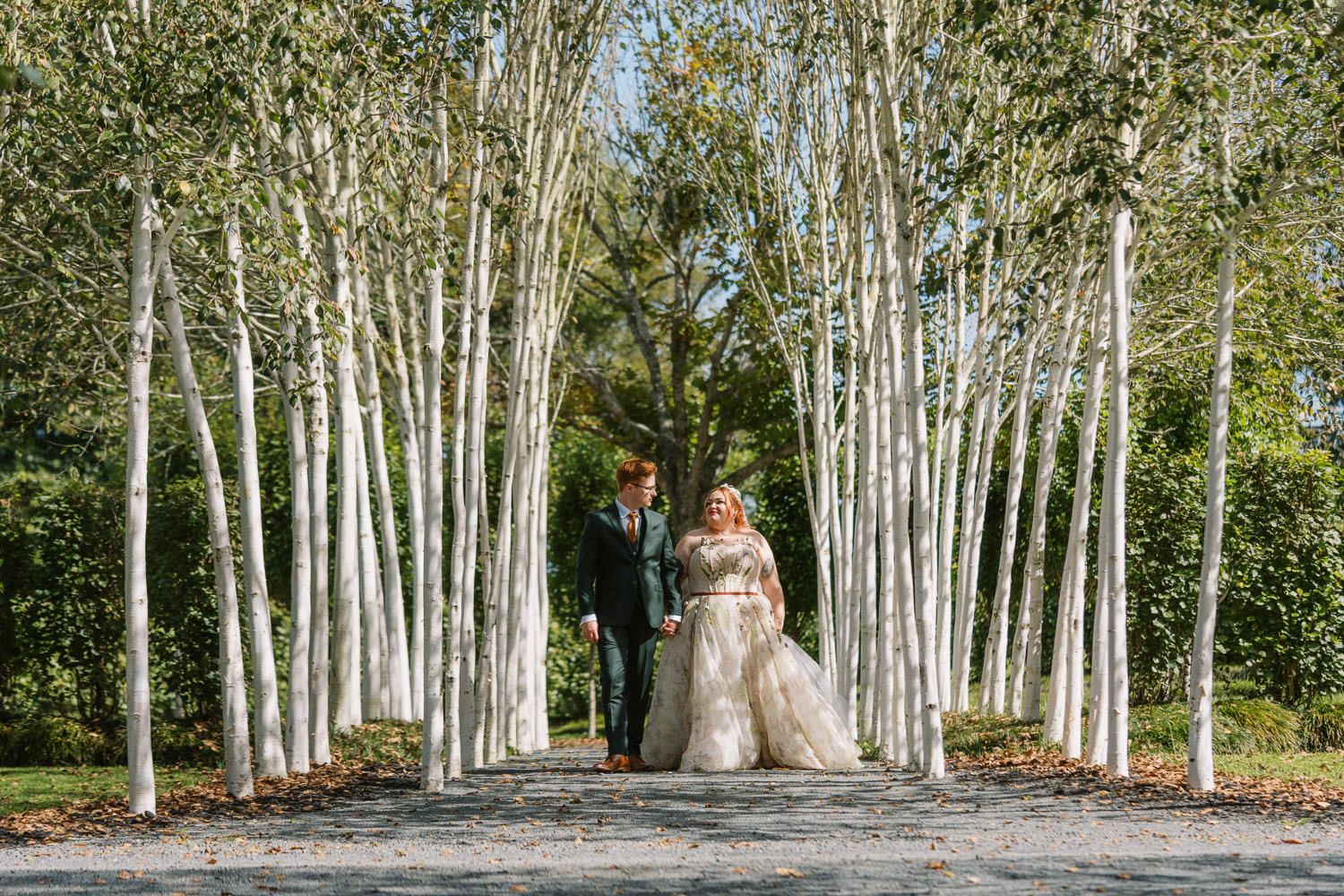 Bride and groom walk hand in hand along a tree-lined path surrounded by white birch trees in a garden setting.