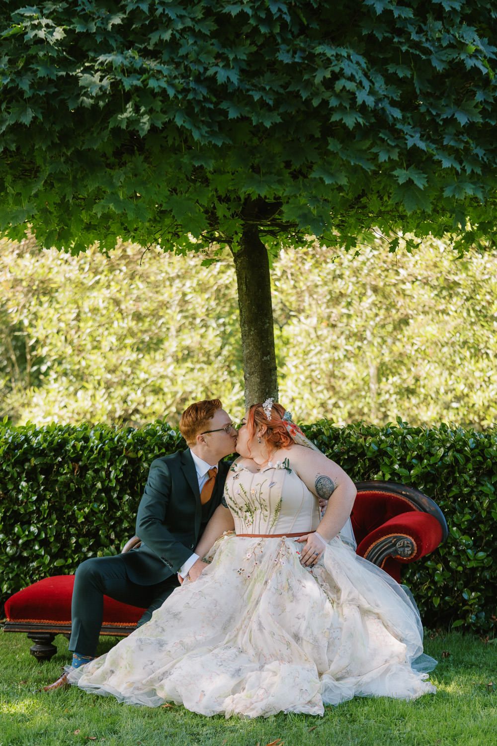 A bride and groom share a romantic kiss on a red velvet sofa in a lush garden wedding setting.