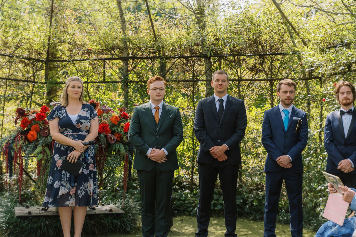 Wedding party stands outdoors under a vine-covered pergola, featuring a woman in floral dress and three men in suits.