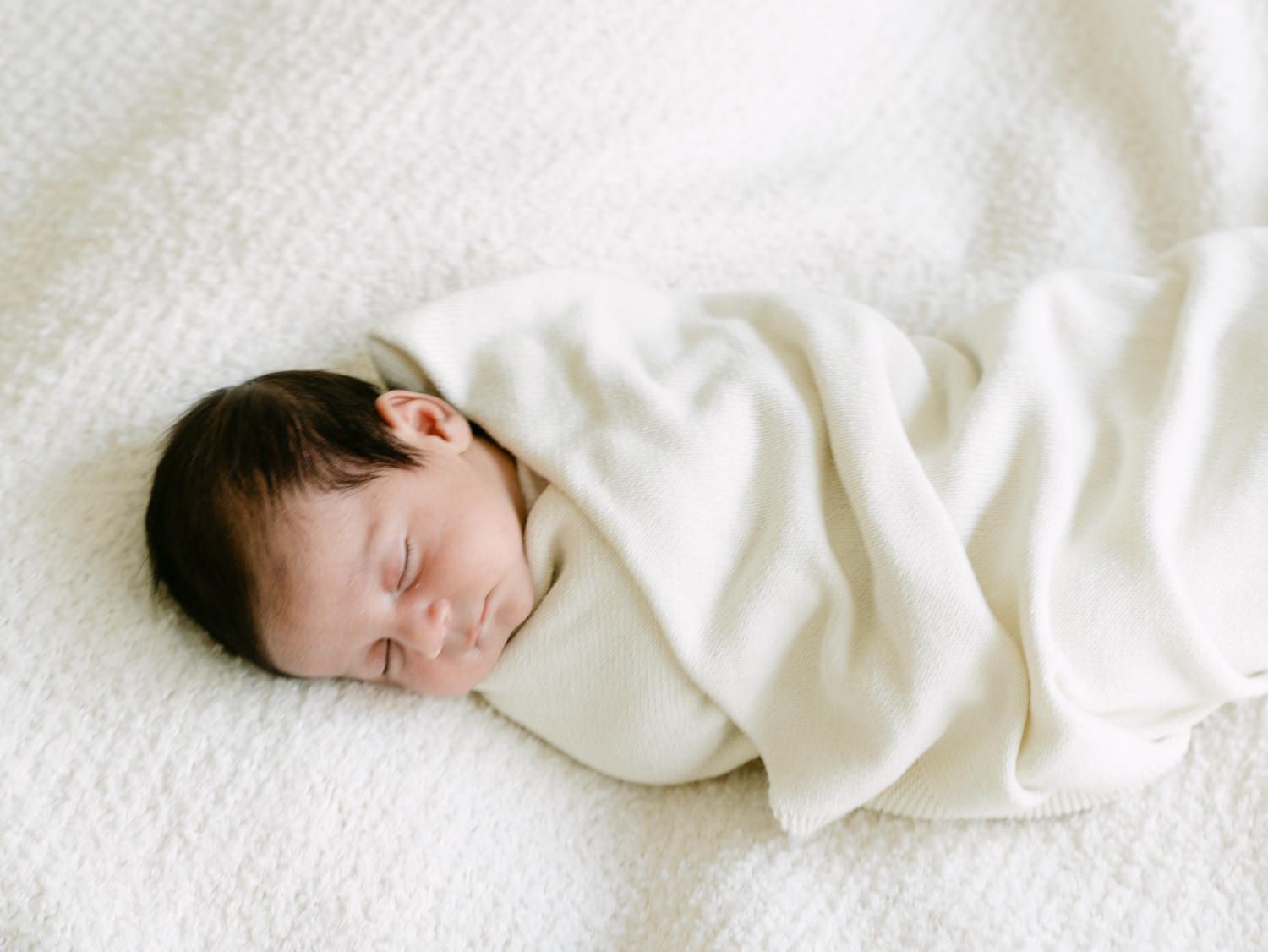 Sleeping swaddled newborn baby wrapped in a cream blanket on white textured surface.
