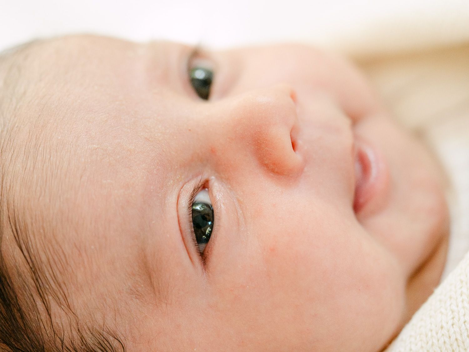 Close-up of alert newborn baby with dark eyes looking upward on soft white surface.