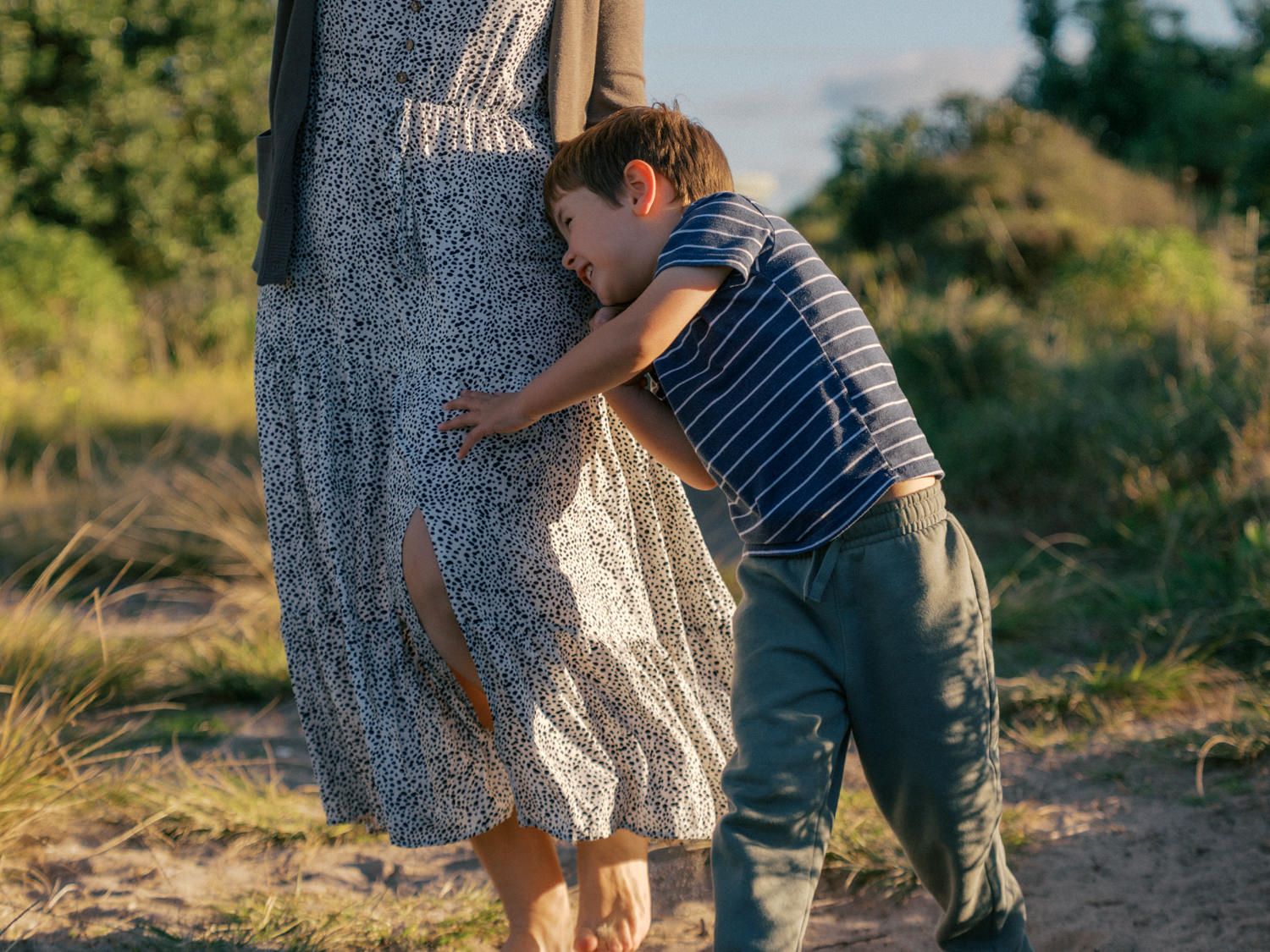 A young boy hugging his mother's legs outdoors in a sunny, natural setting.