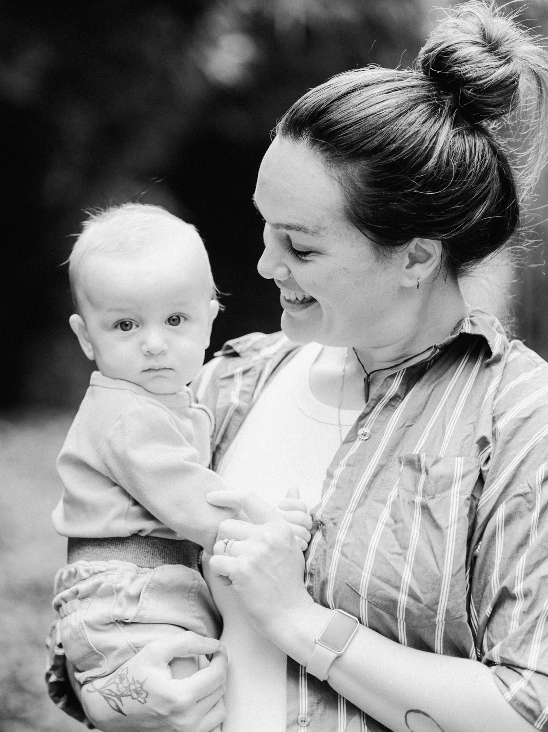 Black and white photo of a smiling mother holding her baby boy outdoors in a garden setting.