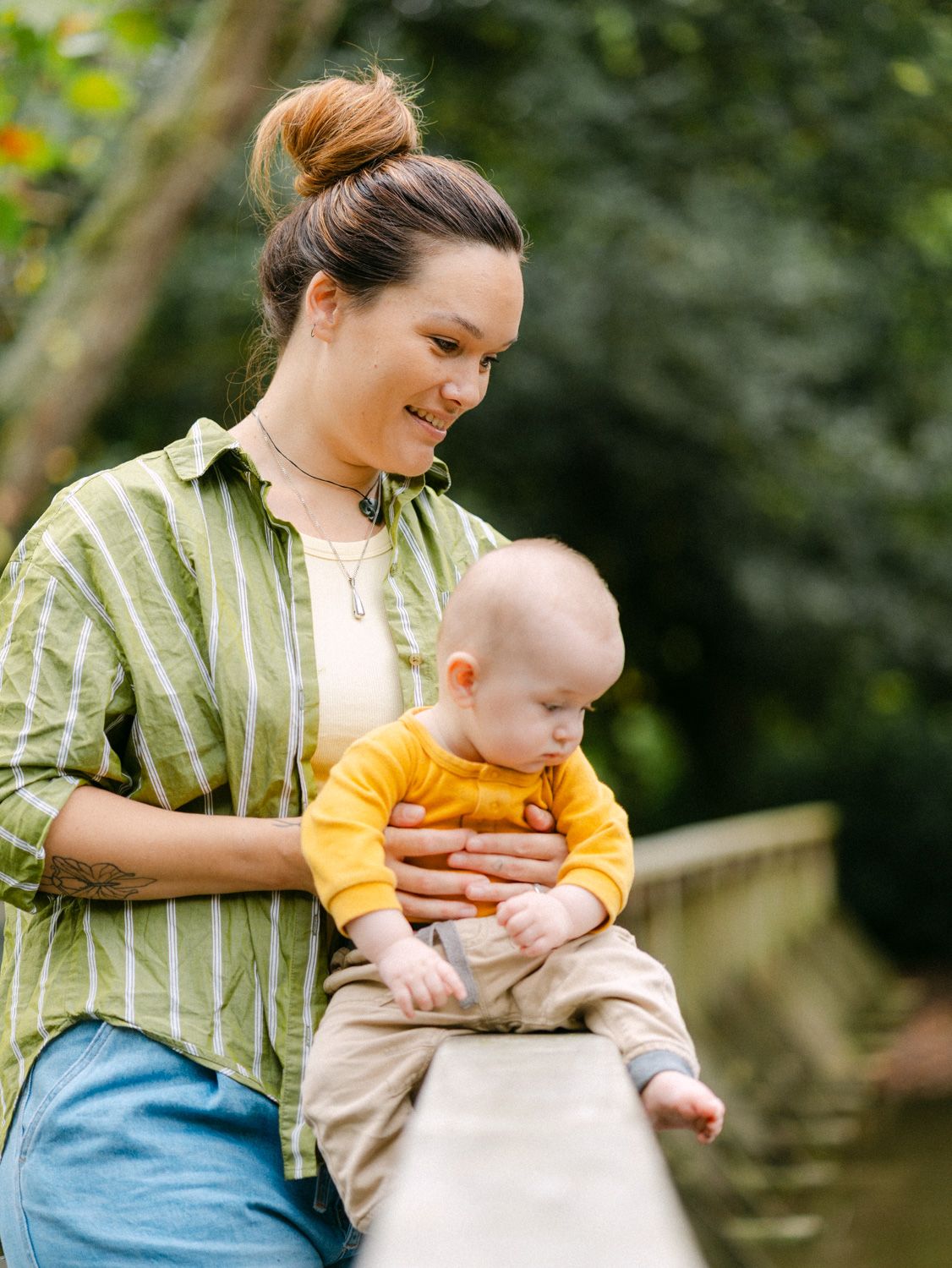 A smiling mother in a green striped shirt holds her baby in a yellow onesie outdoors near a stone bridge.