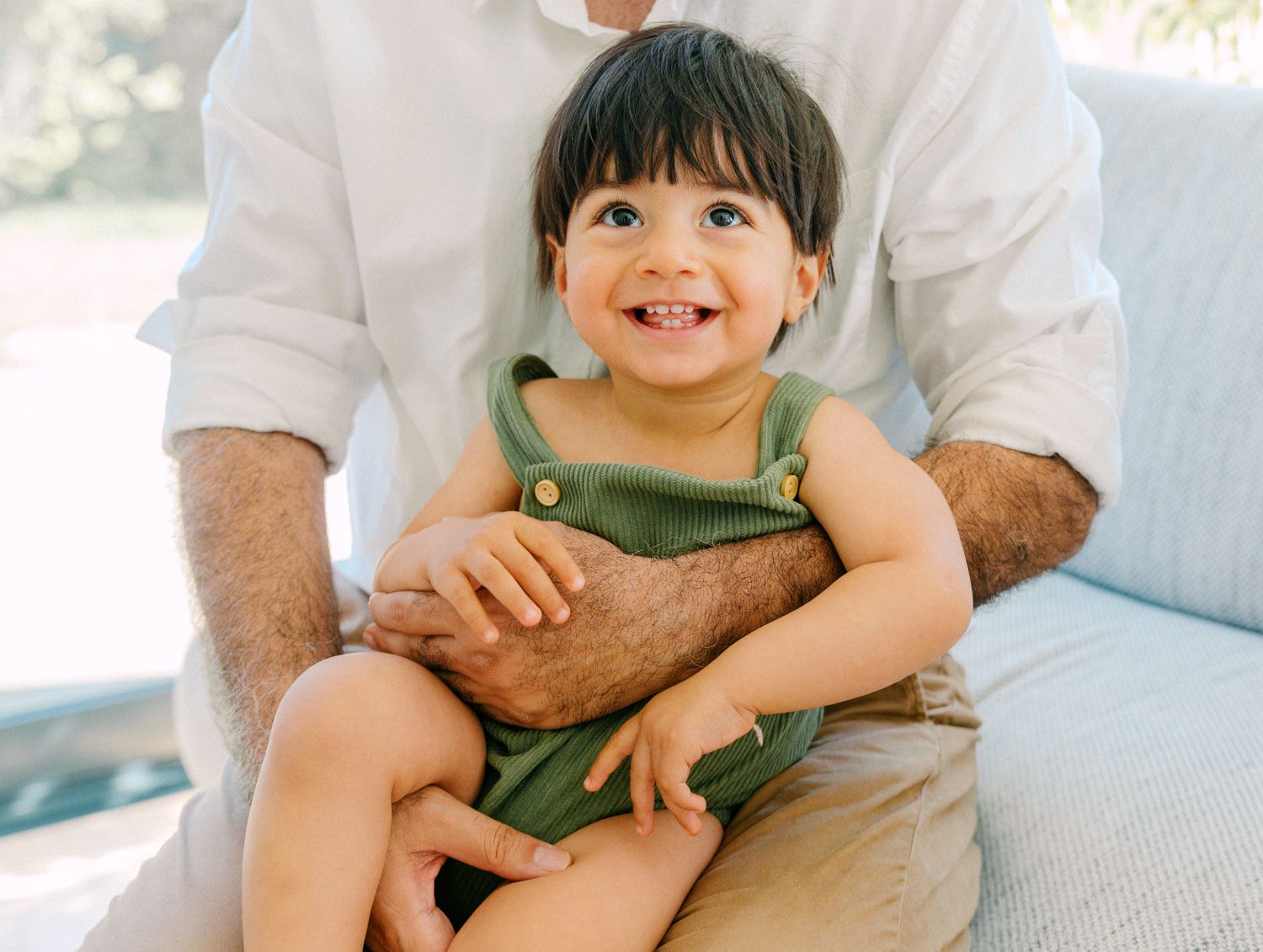 Smiling toddler in green overalls sitting on adult's lap on a light sofa in a bright outdoor setting.