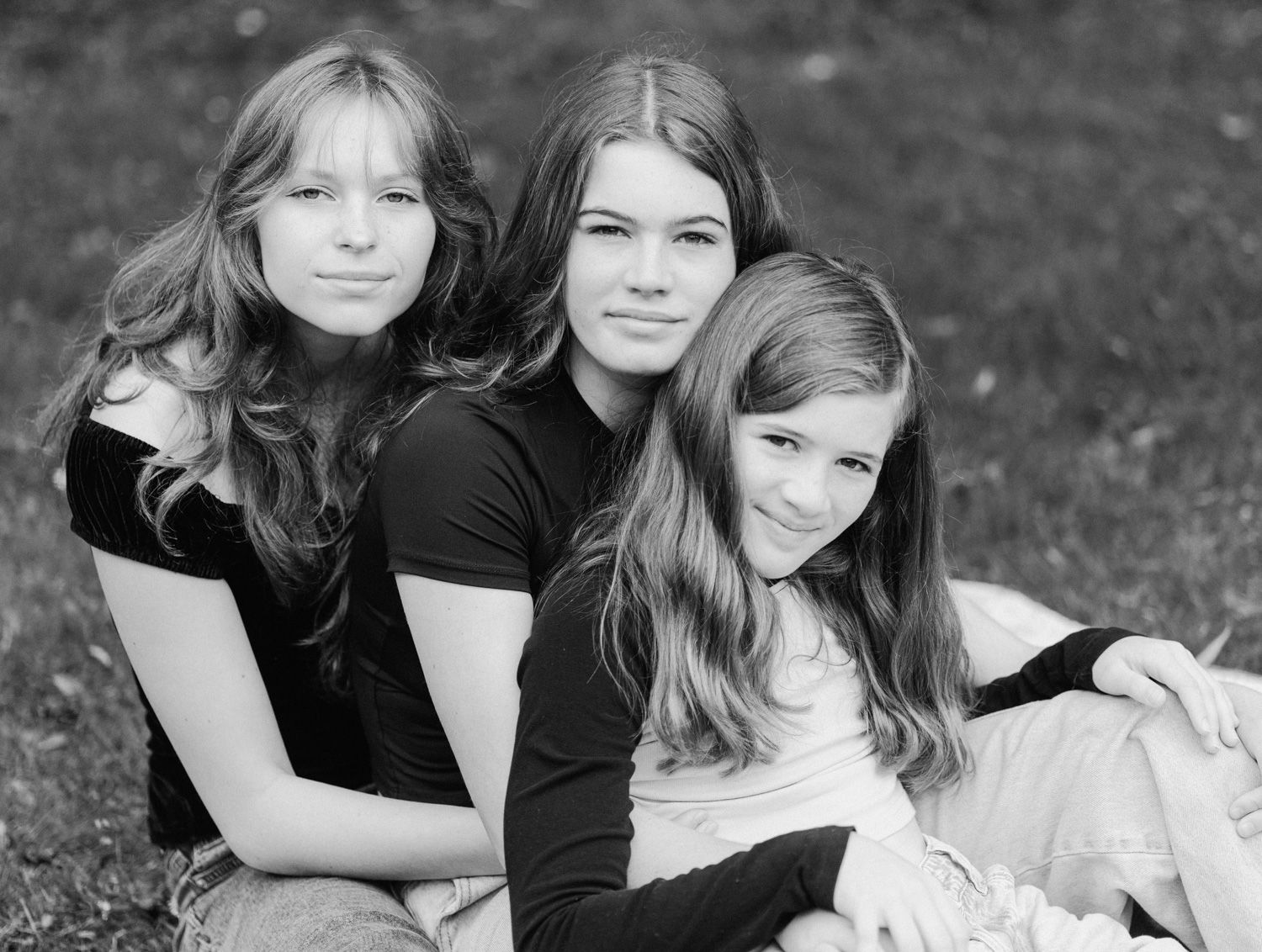 Black and white portrait of three young girls with long hair sitting together outdoors on grass, smiling warmly.