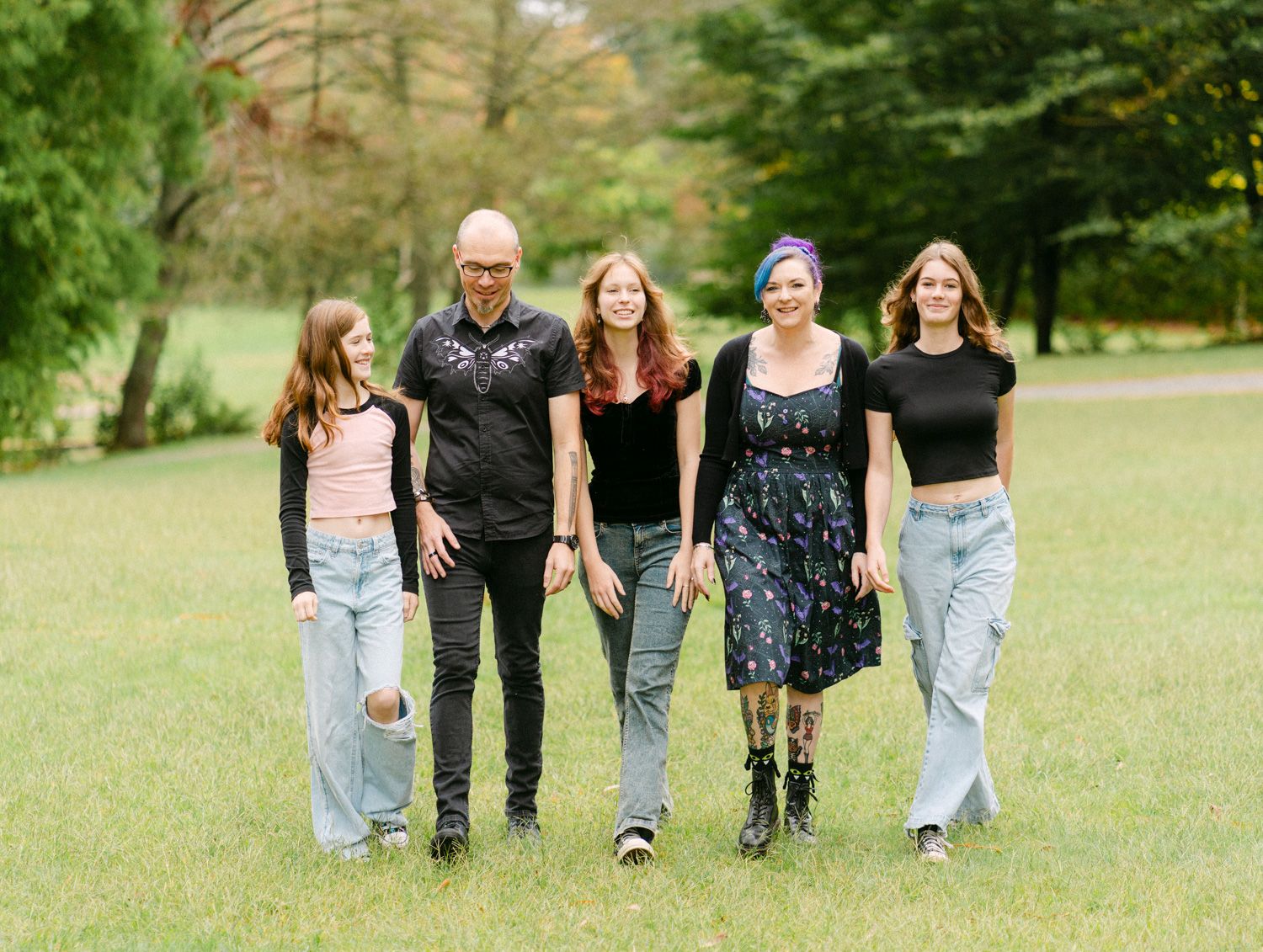 Five people smiling and walking together on green grass in a park, posing for a family portrait outdoors.
