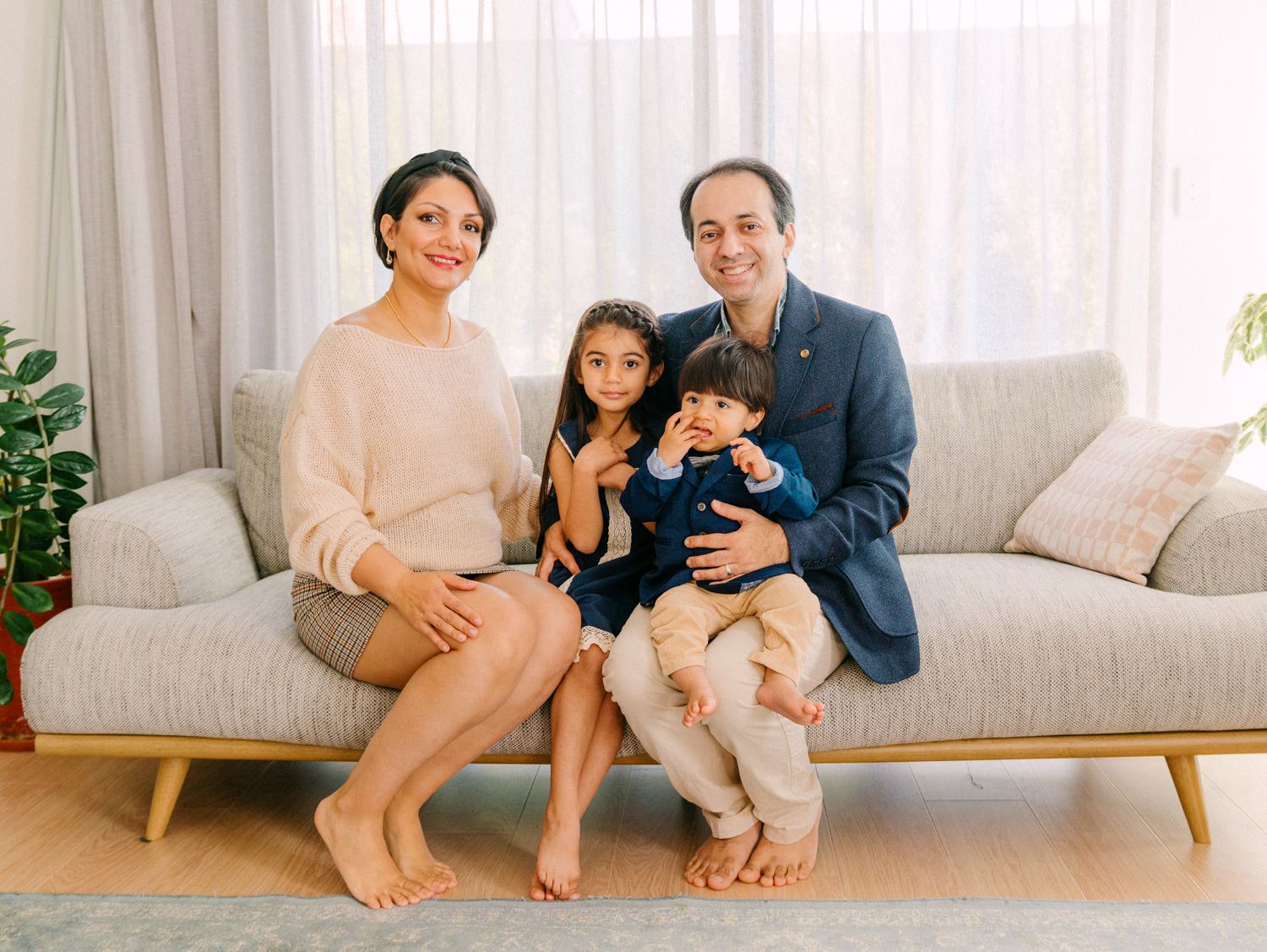 Happy family of four posing together on a gray sofa in a bright living room for a portrait photo.